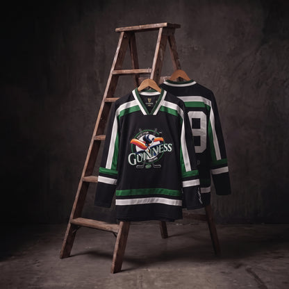 Two Guinness Toucan Hockey Jerseys in black and green are displayed on wooden hangers in front of a wooden ladder, set against a dark, textured background.