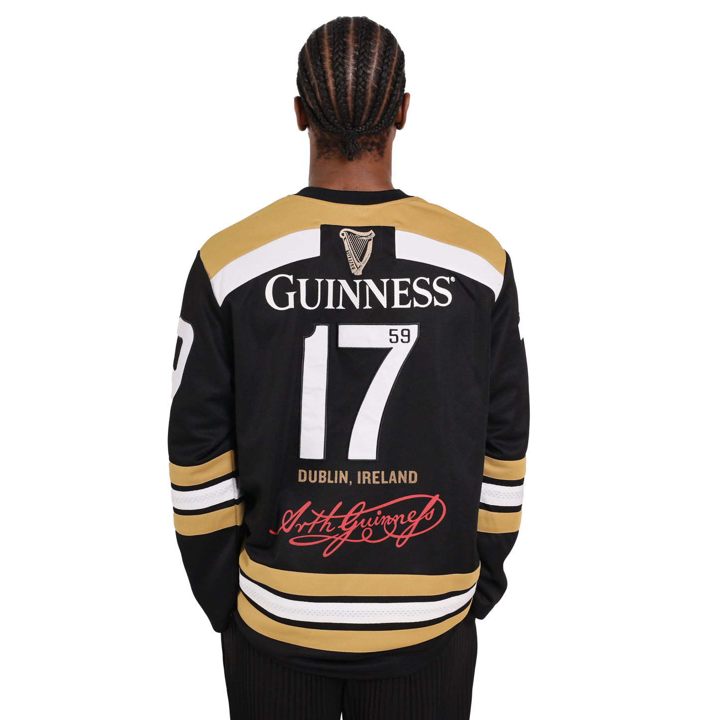 A person with braided hair wearing a black and gold Guinness Webstore US Guinness Tortoise Hockey Jersey, featuring the number 17 and the text "Guinness 1759 Dublin, Ireland" on the back, stands facing away. This sporty design is crafted from recycled polyester for an eco-friendly twist.