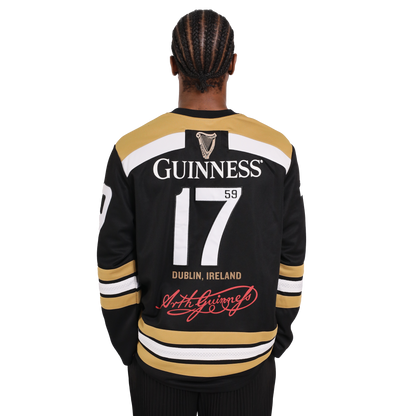 A person with braided hair wearing a black and gold Guinness Webstore US Guinness Tortoise Hockey Jersey, featuring the number 17 and the text "Guinness 1759 Dublin, Ireland" on the back, stands facing away. This sporty design is crafted from recycled polyester for an eco-friendly twist.