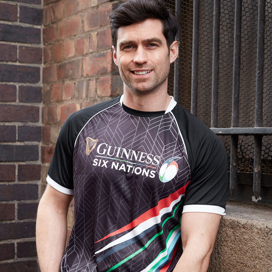 A rugby fan smiling at the camera, wearing a black Guinness Six Nations Performance Tee, standing against a brick wall background.