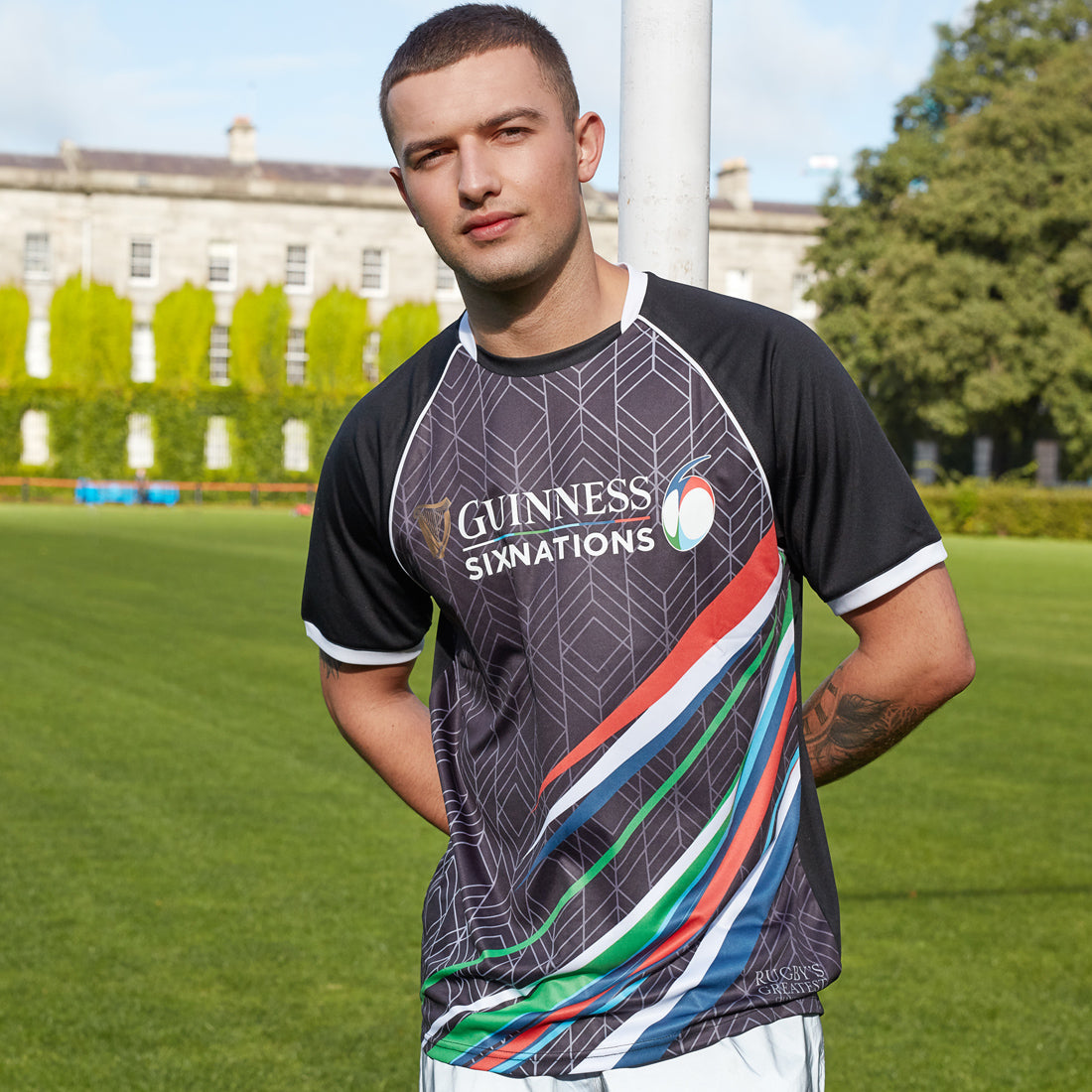 A rugby fan wearing a Guinness Six Nations performance tee stands beside a rugby goal post, with a historical building in the background.