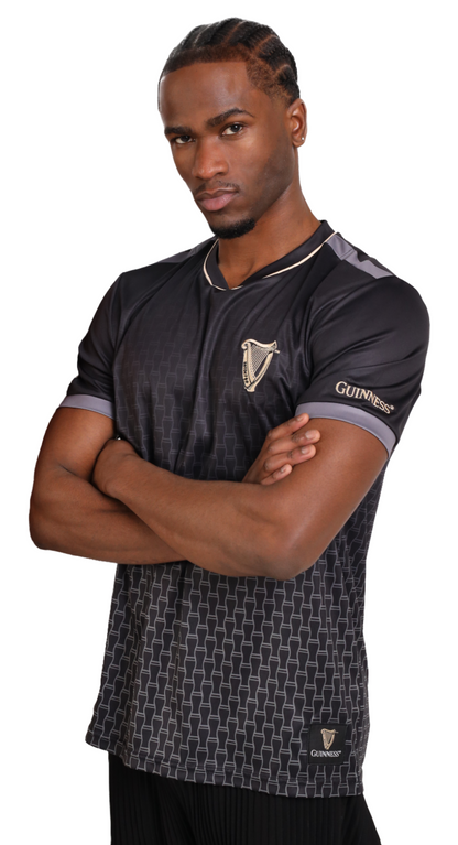 A person stands with arms crossed, wearing a black Guinness Soccer Jersey with a patterned design and the Guinness Webstore US logo on the chest.