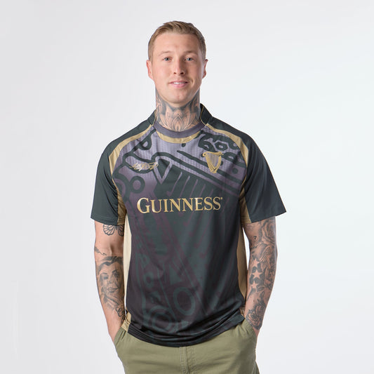 A tattooed individual stands against a plain white background, wearing olive green pants and the Guinness Retro Harp Performance Soccer Jersey by Guinness in black and gold.