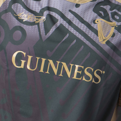 Close-up of the Guinness Retro Harp Performance Soccer Jersey in purple and gold, featuring the Guinness logo, a harp graphic, and a small gold signature on the top left.