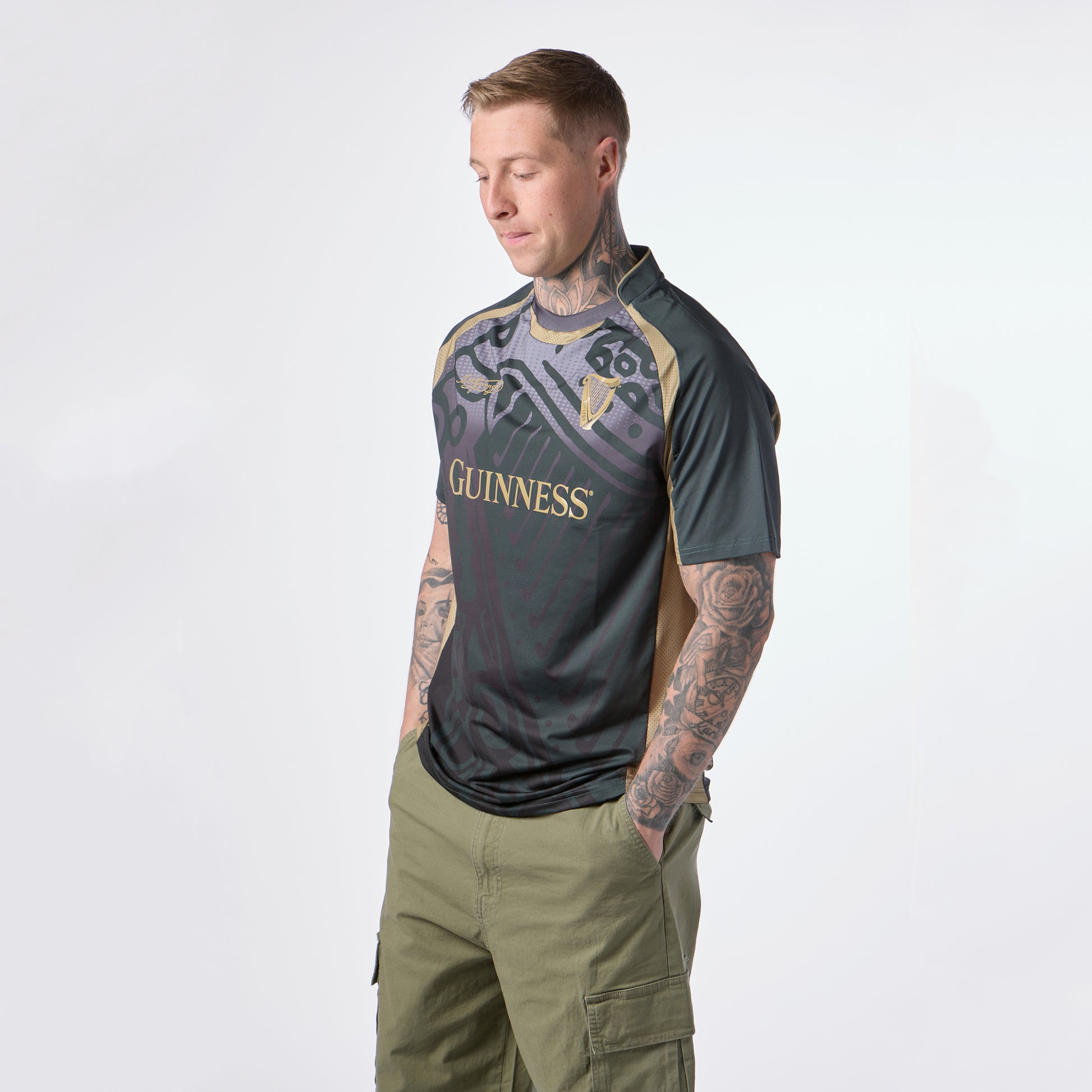 Tattooed man wearing a Guinness Retro Harp Performance Soccer Jersey in green and black, paired with olive green cargo shorts, stands with hands in pockets, looking downward.