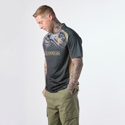 A man with tattoos wearing the Guinness Retro Harp Performance Soccer Jersey and olive cargo pants stands looking down against a plain white background.