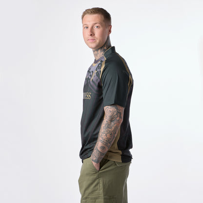 A person with tattoos on their arms stands in profile, wearing a Guinness Retro Harp Performance Soccer Jersey and olive green shorts, hands in pockets, against a plain light background.