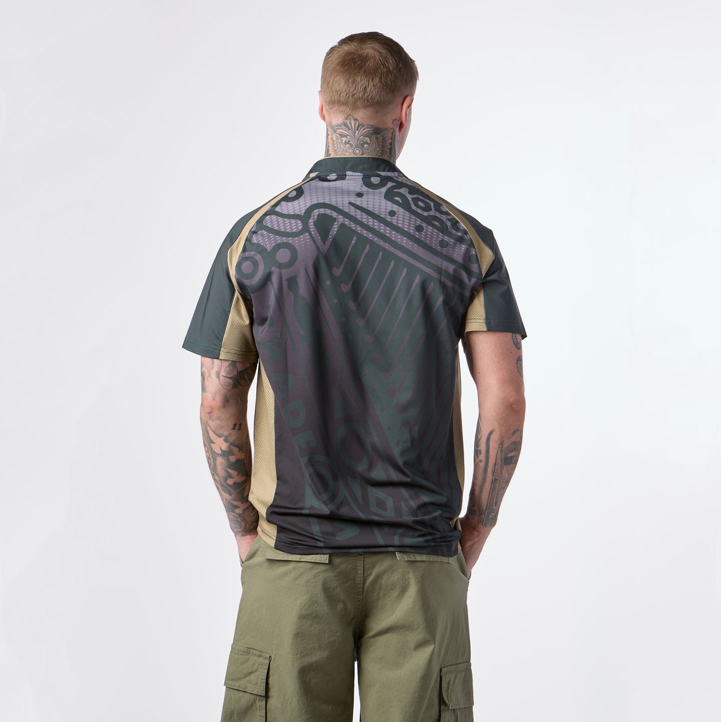 A man with tattoos stands facing away, wearing the Guinness Retro Harp Performance Soccer Jersey—a dark-patterned short-sleeve shirt—paired with olive cargo shorts, against a plain white background.