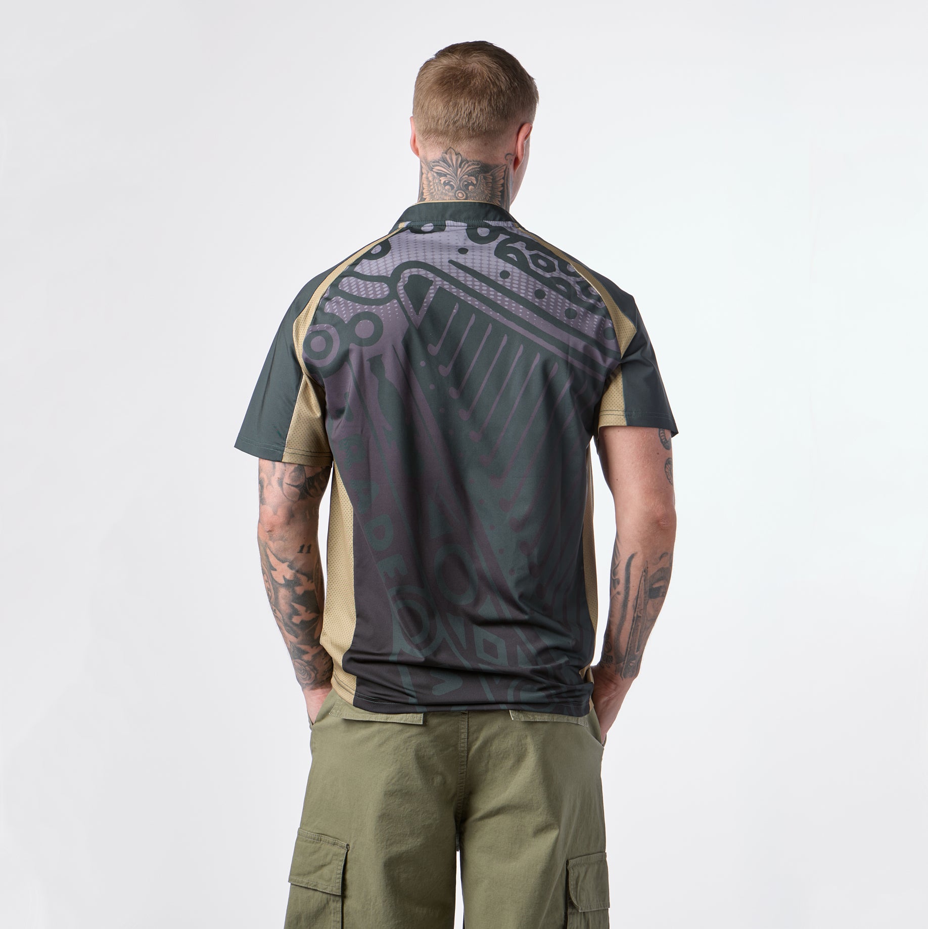 A man with tattoos stands facing away, wearing the Guinness Retro Harp Performance Soccer Jersey—a dark-patterned short-sleeve shirt—paired with olive cargo shorts, against a plain white background.
