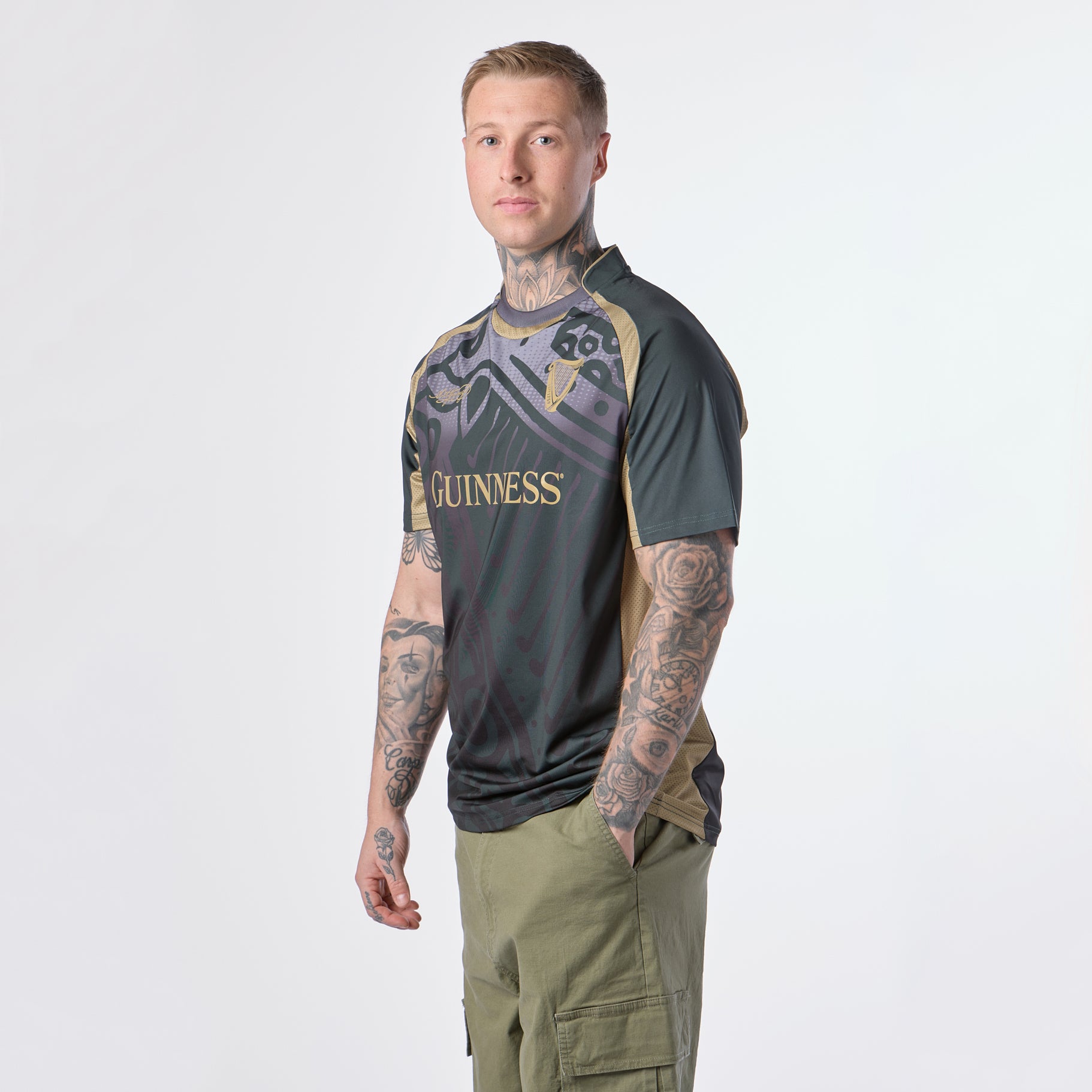 A person with tattoos wearing a black and gold Guinness Retro Harp Performance Soccer Jersey by Guinness and green cargo pants stands facing slightly left against a plain white background.