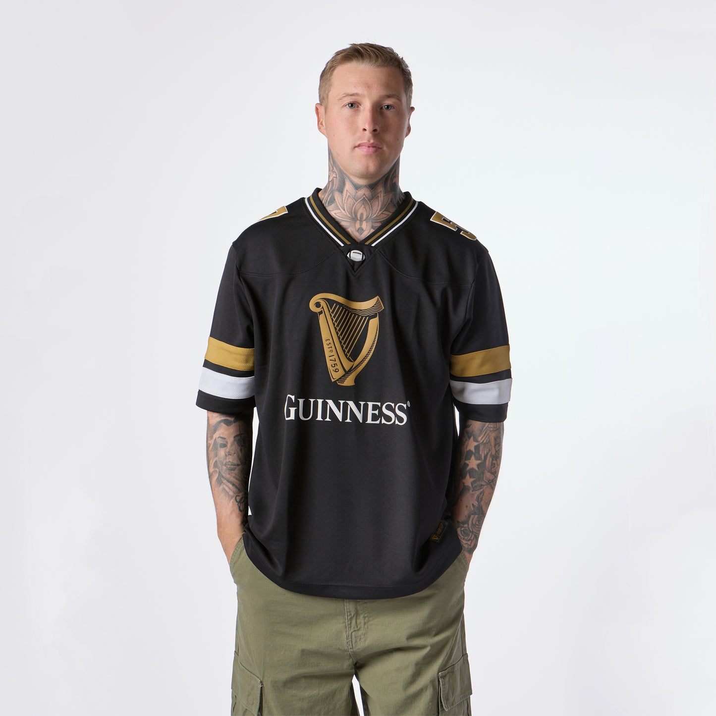 A person with tattoos stands facing forward, wearing the Guinness Black American Football Jersey from Guinness Webstore US and olive green cargo shorts against a plain white background.