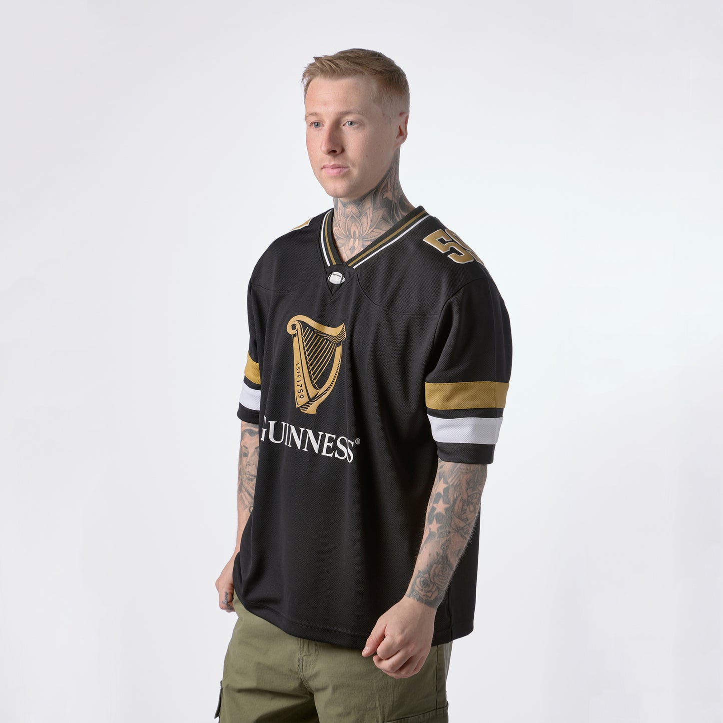 A person with tattoos wears the Guinness Black American Football Jersey from Guinness Webstore US, featuring gold and white stripes, paired with olive green pants, standing against a plain white background.