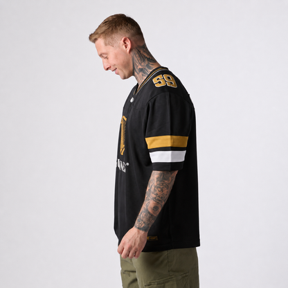 A man with tattoos stands sideways against a plain background, wearing the Guinness Black American Football Jersey from Guinness Webstore US, featuring gold and white stripes and the number 99.