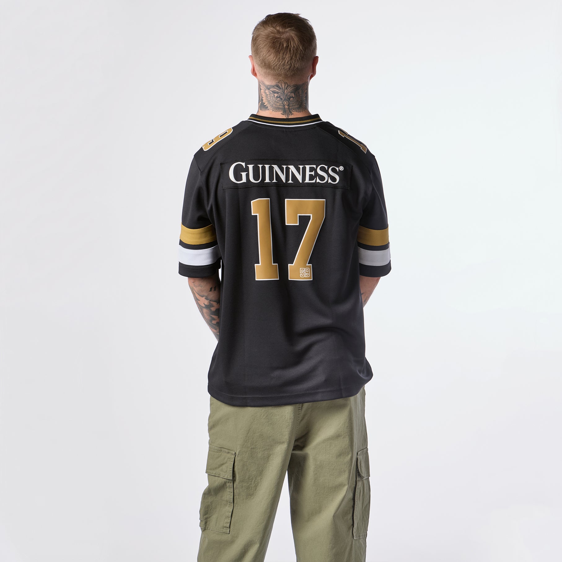 A person with tattoos is seen from behind wearing the Guinness Black American Football Jersey by Guinness Webstore US, featuring “GUINNESS” and the number 17, paired with green cargo pants.