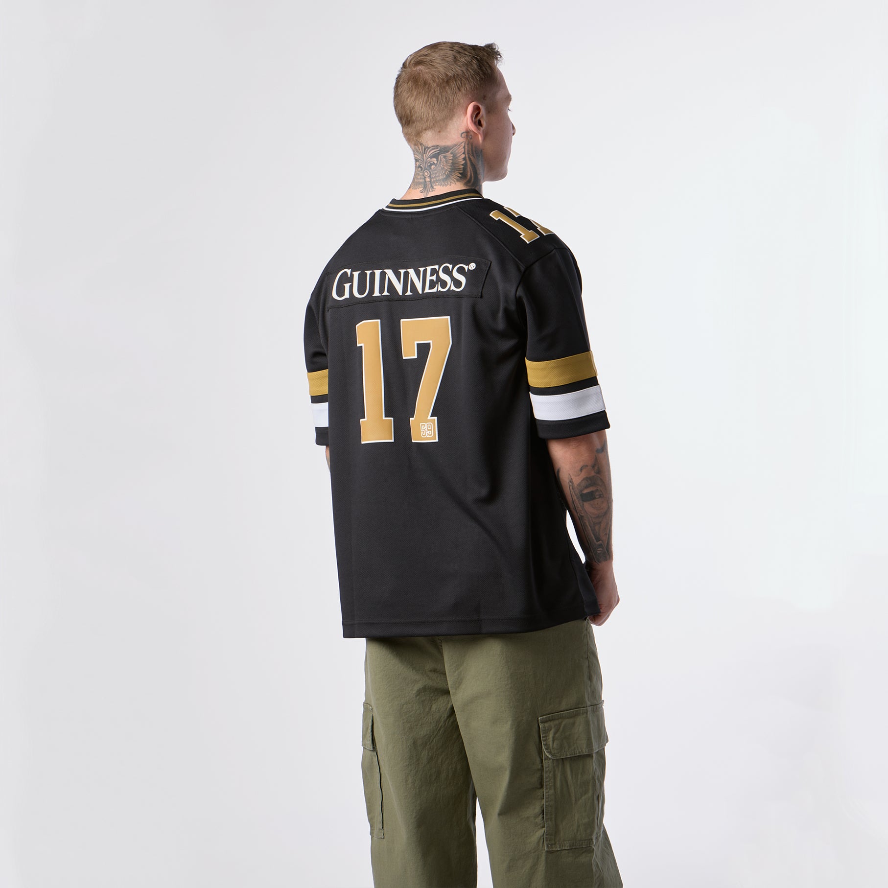 An individual faces away from the camera, dressed in the Guinness Webstore US Guinness Black American Football Jersey—featuring “Guinness 17” and gold and white sleeve stripes—paired with olive green cargo pants, set against a plain white background.