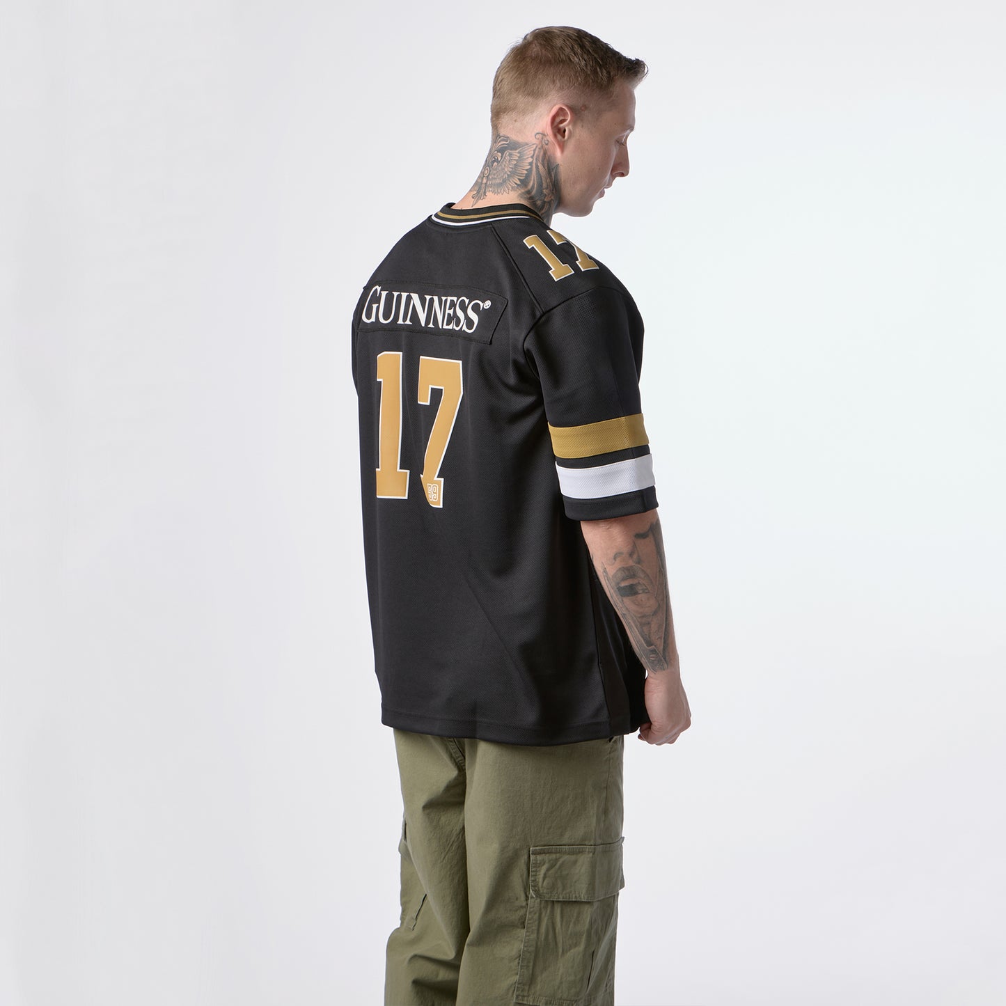 A man stands facing away, wearing the Guinness Black American Football Jersey by Guinness Webstore US with “GUINNESS” and the number 17 in gold lettering, paired with olive green cargo pants.