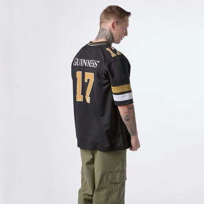 A man stands facing away, wearing the Guinness Black American Football Jersey by Guinness Webstore US with “GUINNESS” and the number 17 in gold lettering, paired with olive green cargo pants.
