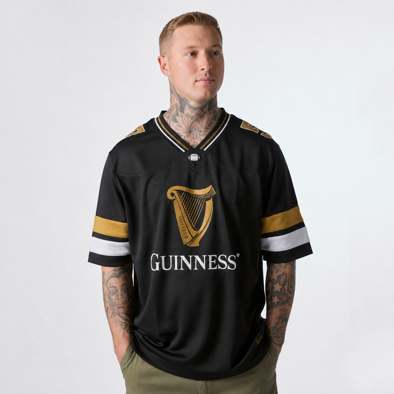 A man with tattoos wears the Guinness Black American Football Jersey from Guinness Webstore US, featuring gold and white stripes, while standing against a plain light background.
