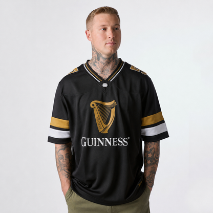 A man with tattoos wears the Guinness Black American Football Jersey from Guinness Webstore US, featuring gold and white stripes, while standing against a plain light background.