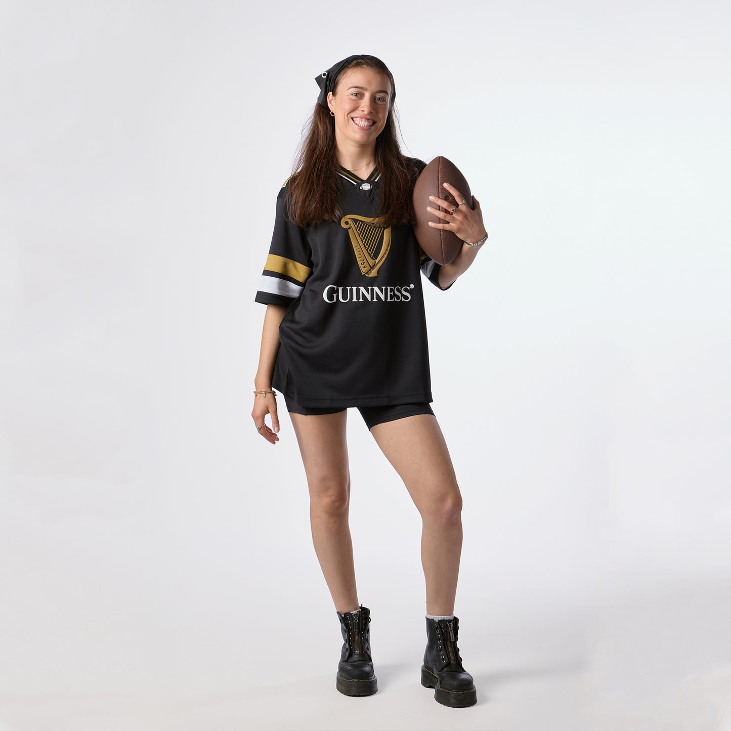 A woman wearing the Guinness Black American Football Jersey from Guinness Webstore US smiles and holds an American football, standing against a plain white background to proudly showcase her love for Guinness apparel.
