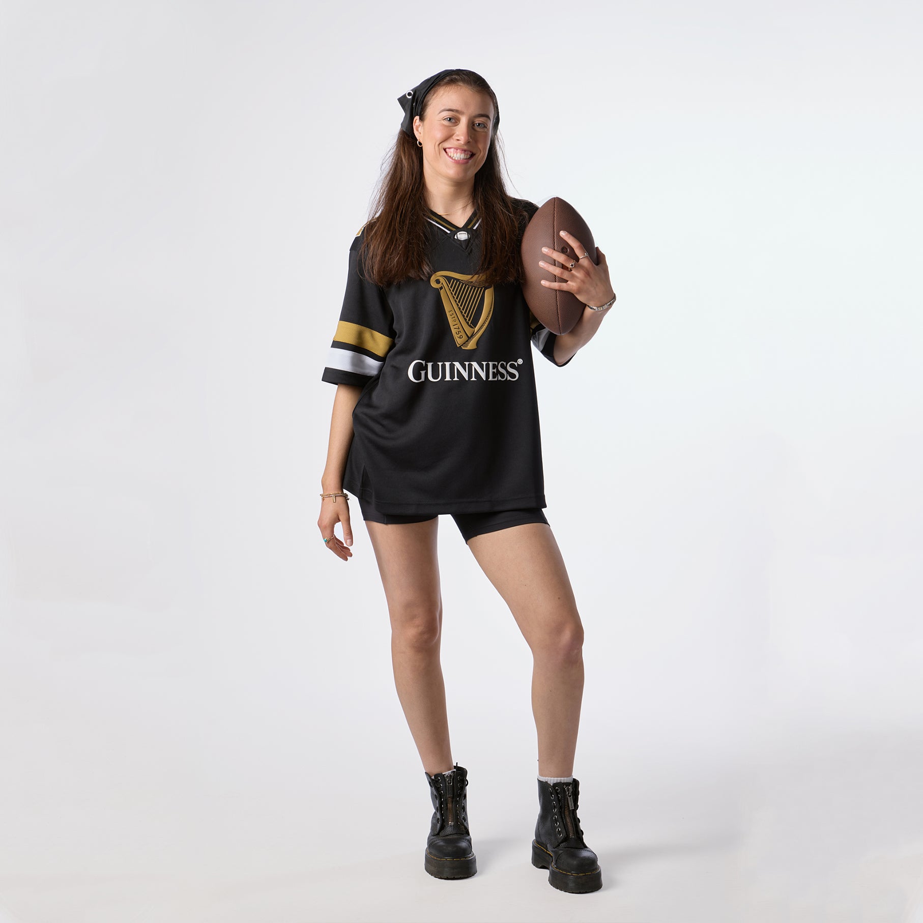A woman wearing the Guinness Black American Football Jersey from Guinness Webstore US smiles and holds an American football, standing against a plain white background to proudly showcase her love for Guinness apparel.