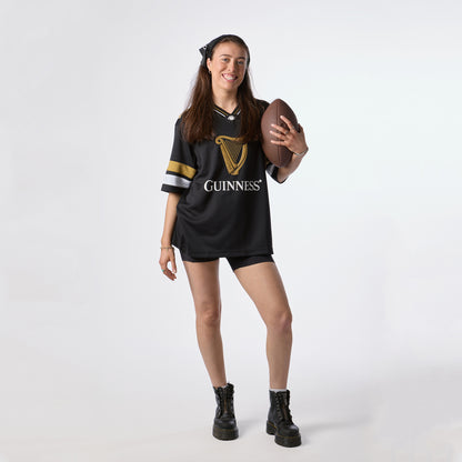 A woman wearing the Guinness Black American Football Jersey from Guinness Webstore US smiles and holds an American football, standing against a plain white background to proudly showcase her love for Guinness apparel.
