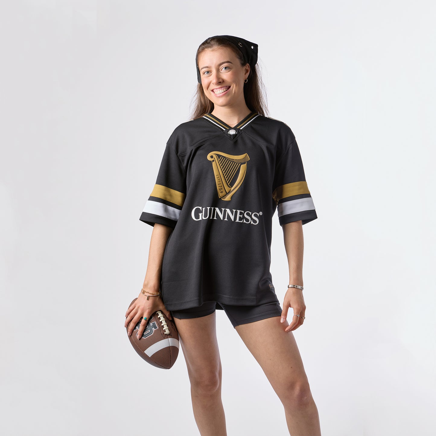 A woman stands and smiles against a plain white background, holding a football in one hand while wearing the Guinness Black American Football Jersey from Guinness Webstore US and matching shorts.