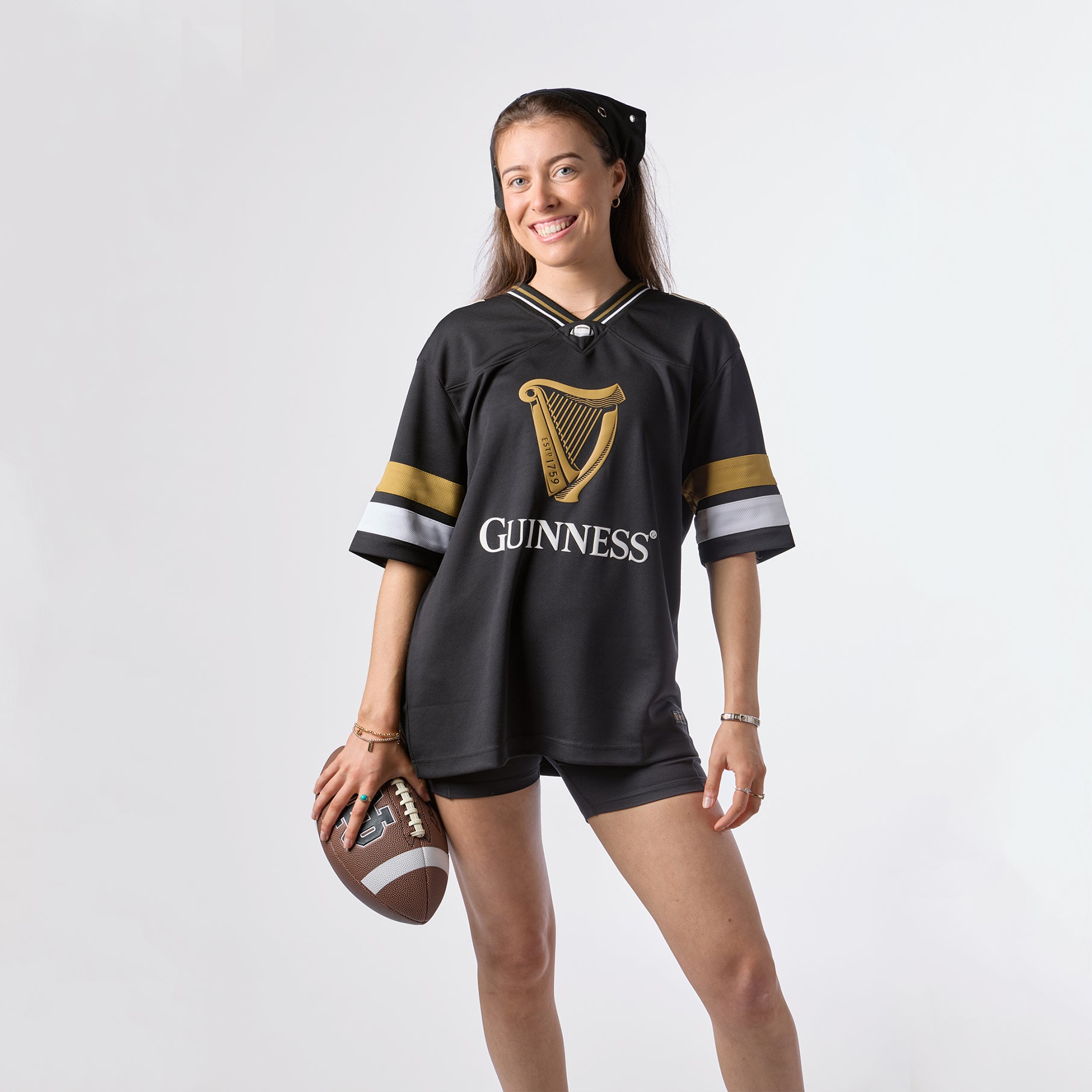 A woman stands and smiles against a plain white background, holding a football in one hand while wearing the Guinness Black American Football Jersey from Guinness Webstore US and matching shorts.