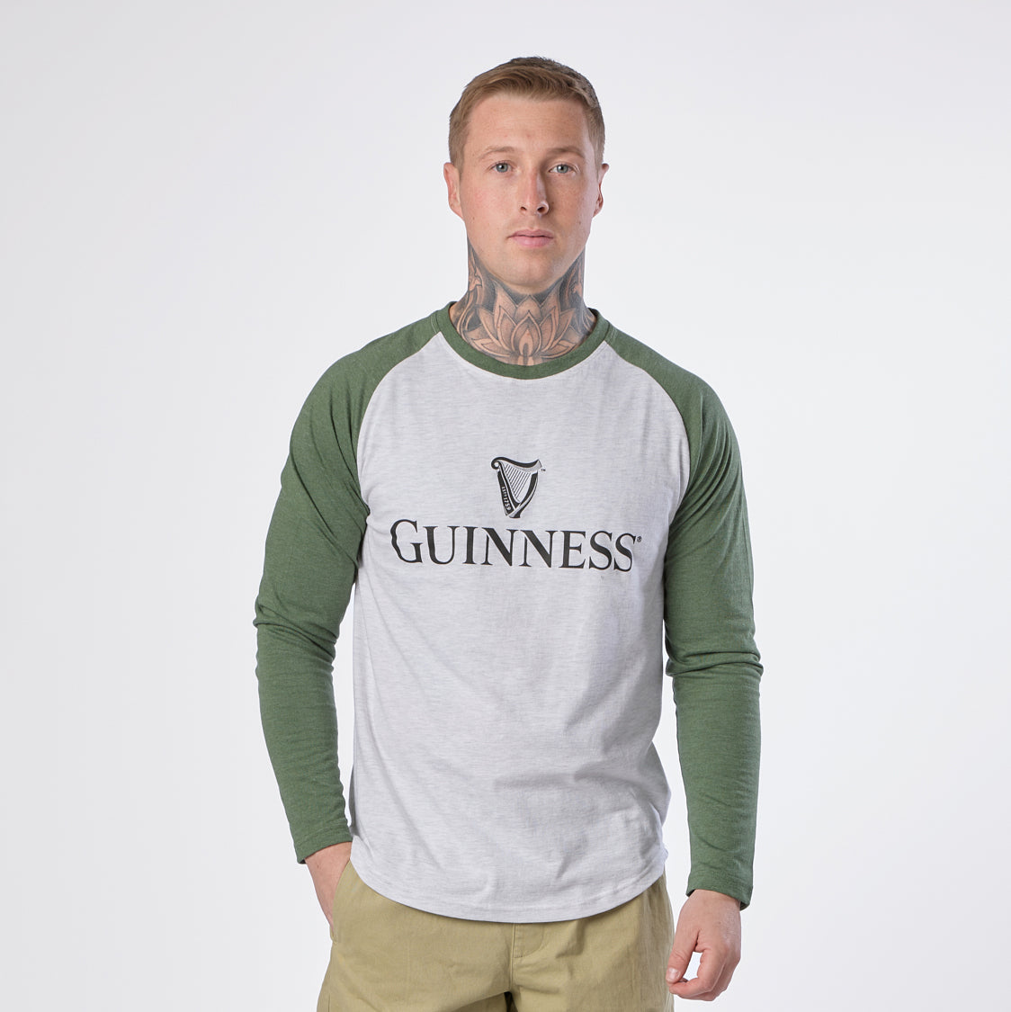 A man wears the Guinness Baseball T-Shirt by Guinness in heathered grey green, featuring the signature “Guinness” logo, standing against a plain white background with his hands in his pockets.