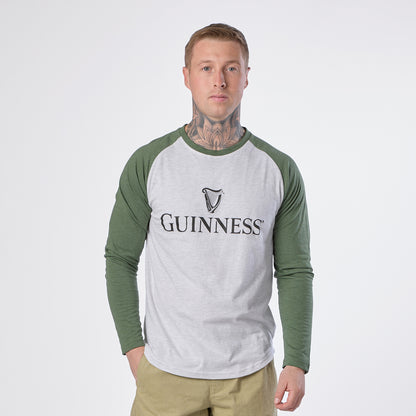 A man wears the Guinness Baseball T-Shirt by Guinness in heathered grey green, featuring the signature “Guinness” logo, standing against a plain white background with his hands in his pockets.