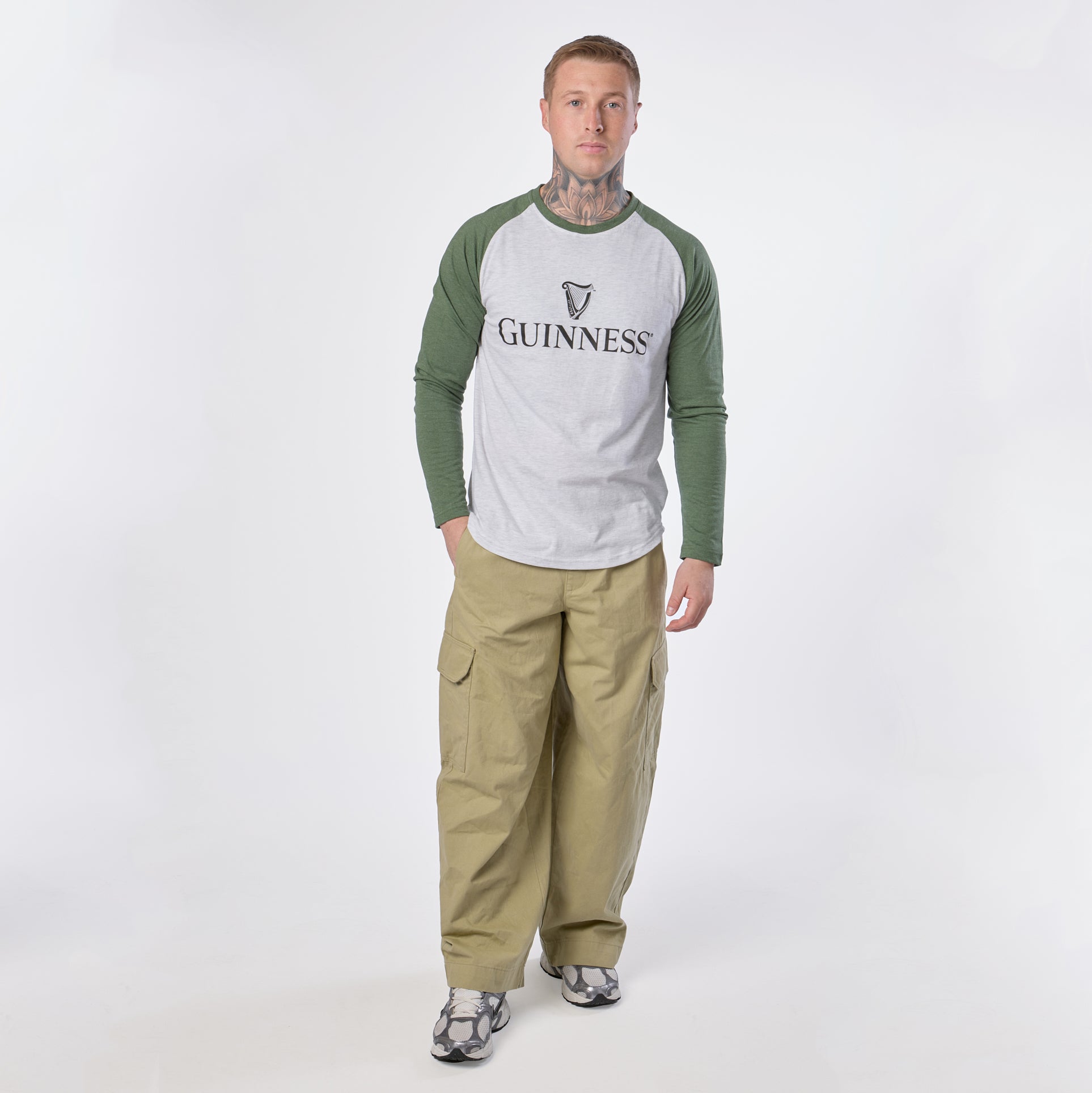 A man stands facing forward against a white background, wearing a Guinness Baseball T-Shirt in heathered grey green with green sleeves, paired with khaki cargo pants and gray sneakers.