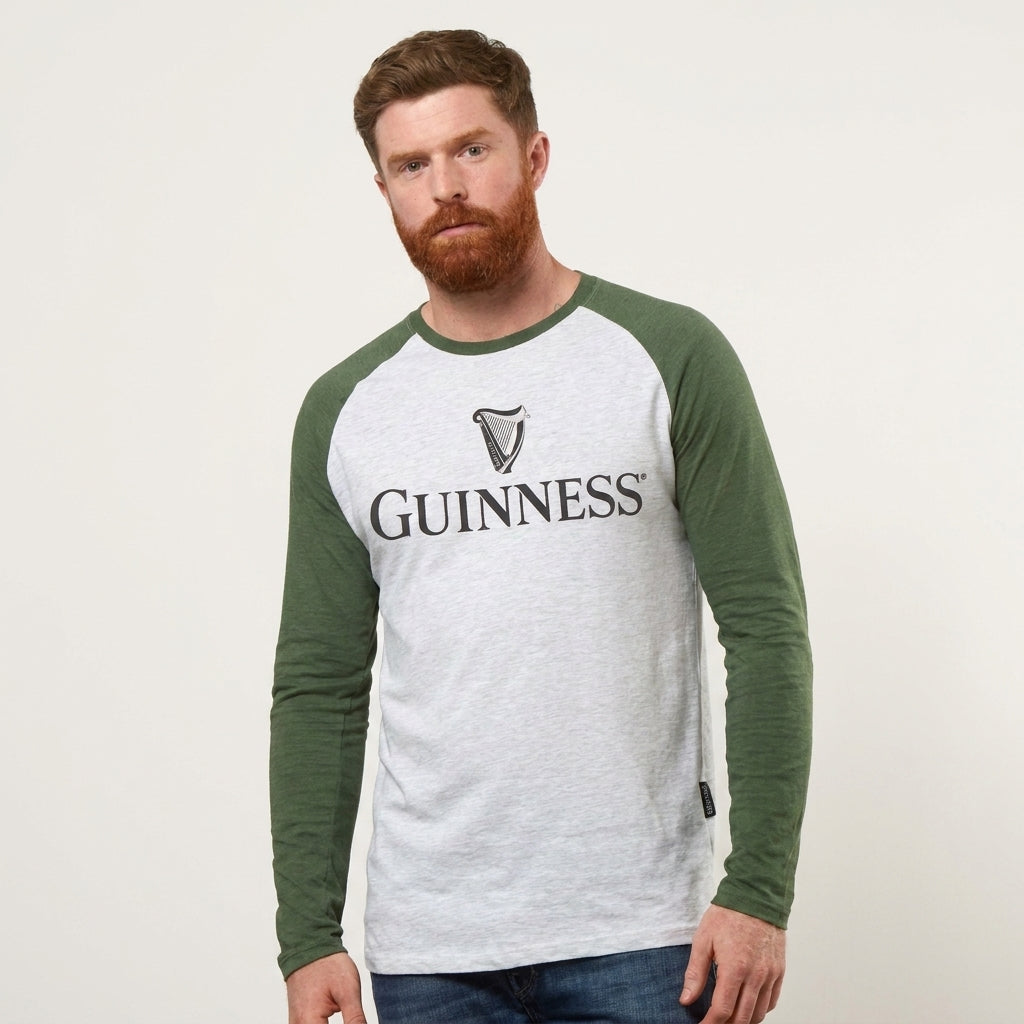 Man with a beard wearing a Guinness Baseball T-Shirt by Guinness, featuring white and green raglan sleeves, stands against a plain light background.