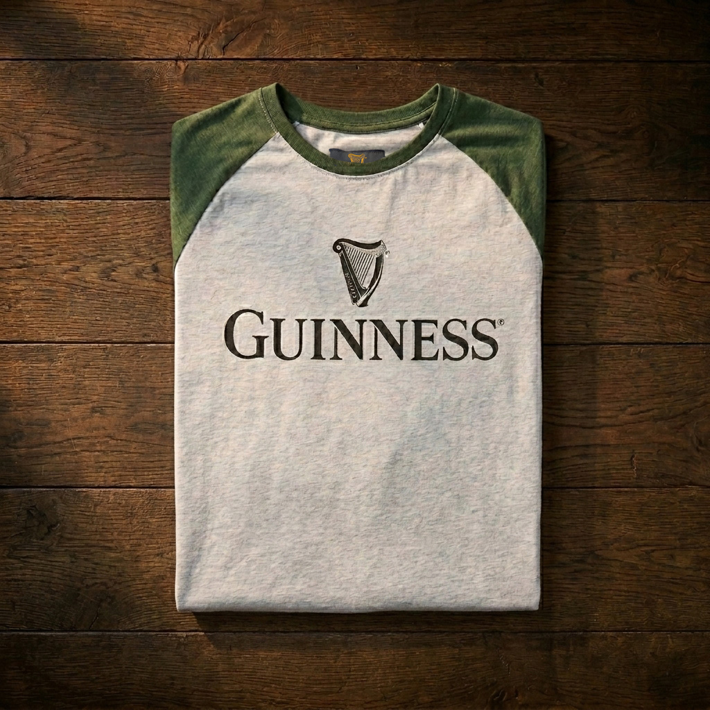 The Guinness Baseball T-Shirt by Guinness features gray and green raglan sleeves, a front logo with harp emblem, and soft heathered jersey cotton, displayed on a wooden surface.