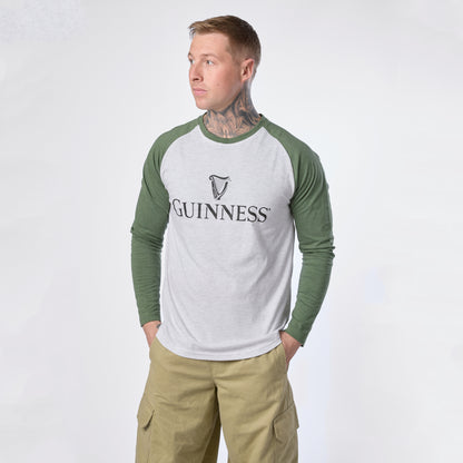 A man stands with his hands in his pockets against a plain background, wearing khaki pants and a white and green Guinness Baseball T-Shirt by Guinness.