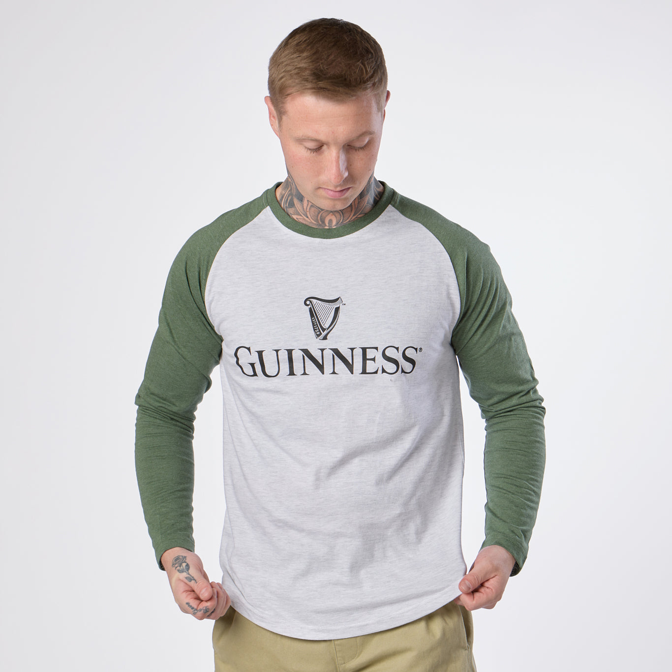 A person with short hair and visible neck tattoos wears the Guinness Baseball T-Shirt by Guinness, featuring heathered grey and green long sleeves and a bold “Guinness” logo on the front. They look down at the shirt, perfectly showcasing this ideal choice for baseball lovers.