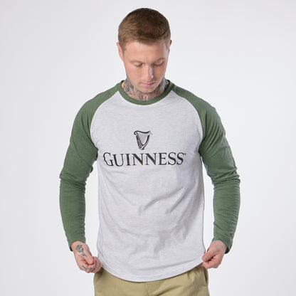 A person with short hair and visible neck tattoos wears the Guinness Baseball T-Shirt by Guinness, featuring heathered grey and green long sleeves and a bold “Guinness” logo on the front. They look down at the shirt, perfectly showcasing this ideal choice for baseball lovers.