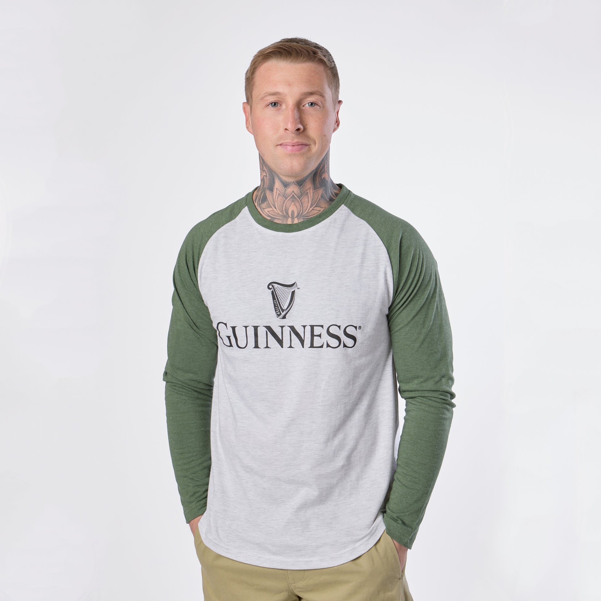 A person with tattoos on their neck is standing against a plain white background, wearing the Guinness Baseball T-Shirt—a white and green long-sleeve shirt from Guinness—paired with light tan pants.