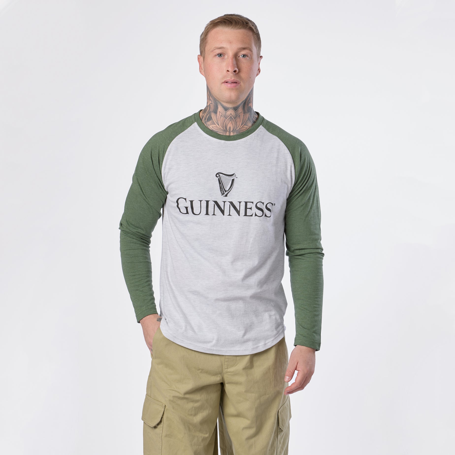 A man with tattoos wears the Guinness Baseball T-Shirt by Guinness in white and green along with khaki pants, standing against a plain white background and looking directly at the camera.