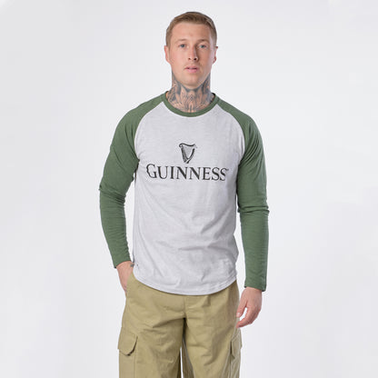 A man with tattoos wears the Guinness Baseball T-Shirt by Guinness in white and green along with khaki pants, standing against a plain white background and looking directly at the camera.