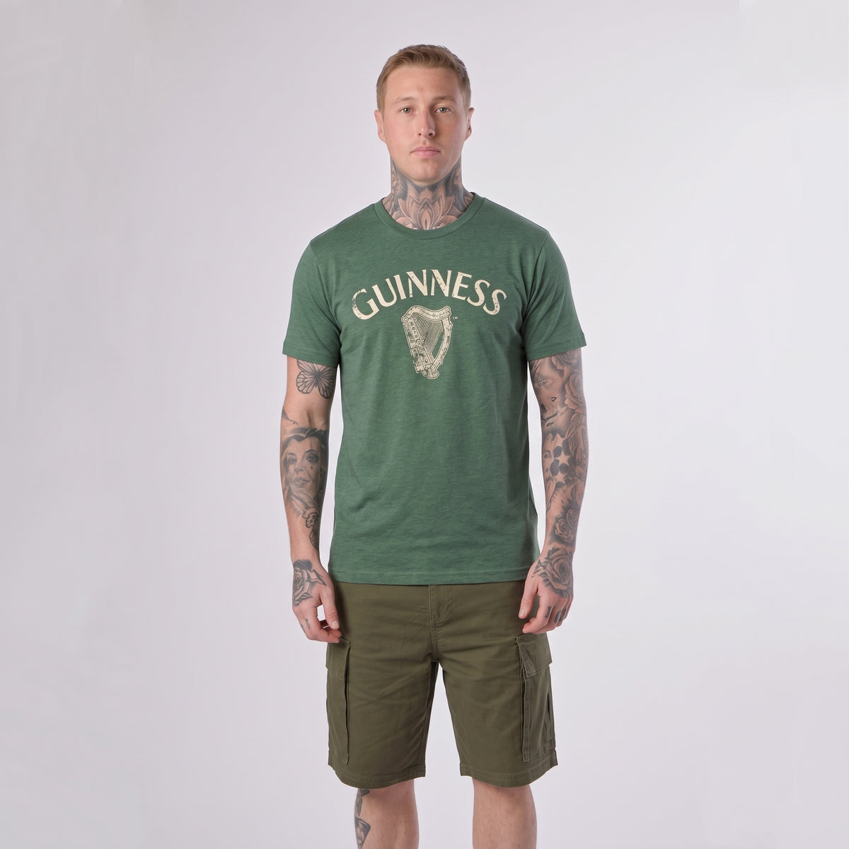 A person with tattoos wears a green Guinness Vintage Harp Tee by Guinness and olive cargo shorts, standing against a plain light background.