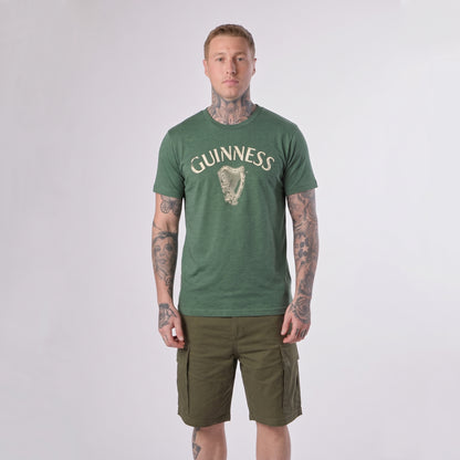 A person with tattoos wears a green Guinness Vintage Harp Tee by Guinness and olive cargo shorts, standing against a plain light background.
