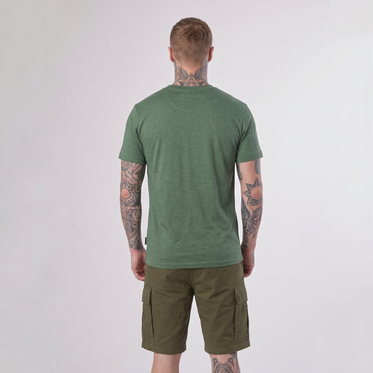 A man with tattoos stands facing away, wearing a green Guinness Vintage Harp Tee by Guinness and olive cargo shorts against a plain light background.