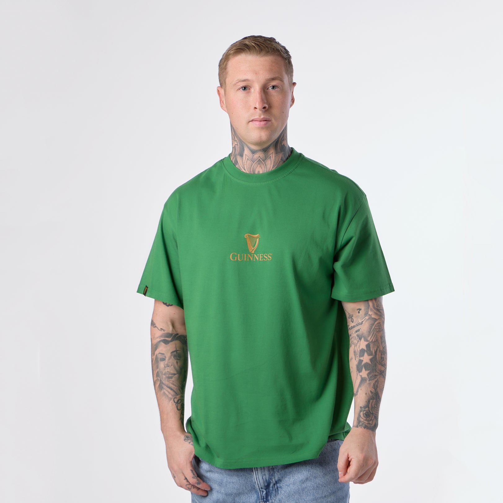 A man stands facing forward, wearing the Guinness Gilroy Toucan Tee in green from the Guinness Webstore US and blue jeans against a plain light background.