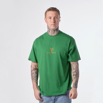 A man stands facing forward, wearing the Guinness Gilroy Toucan Tee in green from the Guinness Webstore US and blue jeans against a plain light background.