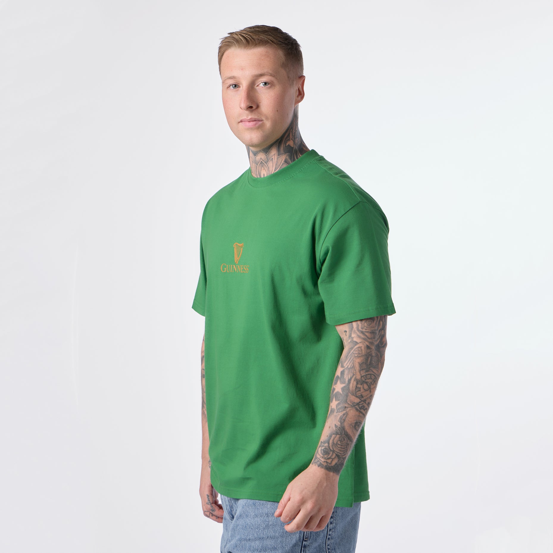 A man with tattoos, wearing the Guinness Gilroy Toucan Tee in green from Guinness Webstore US and blue jeans, stands facing slightly to the side against a plain white background.