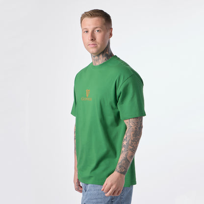 A man with tattoos, wearing the Guinness Gilroy Toucan Tee in green from Guinness Webstore US and blue jeans, stands facing slightly to the side against a plain white background.