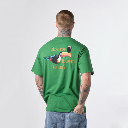 A tattooed man wearing the Guinness Gilroy Toucan Tee – Green from Guinness Webstore US, paired with jeans, stands with his back to the camera. The boxy shirt, part of the Gilroy collection, displays a Guinness toucan, a pint, and the phrase “Are you Ibuoan to me?”.