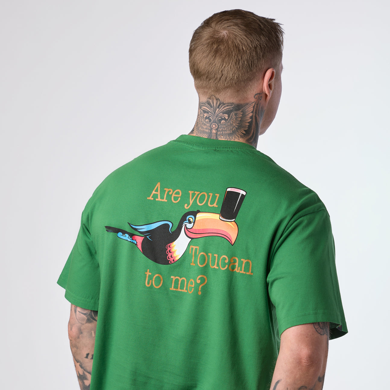 A person with tattoos wears the Guinness Gilroy Toucan Tee – Green from the Guinness Webstore US, which features a cartoon Guinness toucan holding a pint and the text “Are you Toucan to me?” on the back.