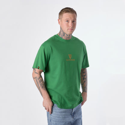 Wearing the Guinness Gilroy Toucan Tee – Green from Guinness Webstore US and light blue jeans, a person with tattoos stands against a plain white background.