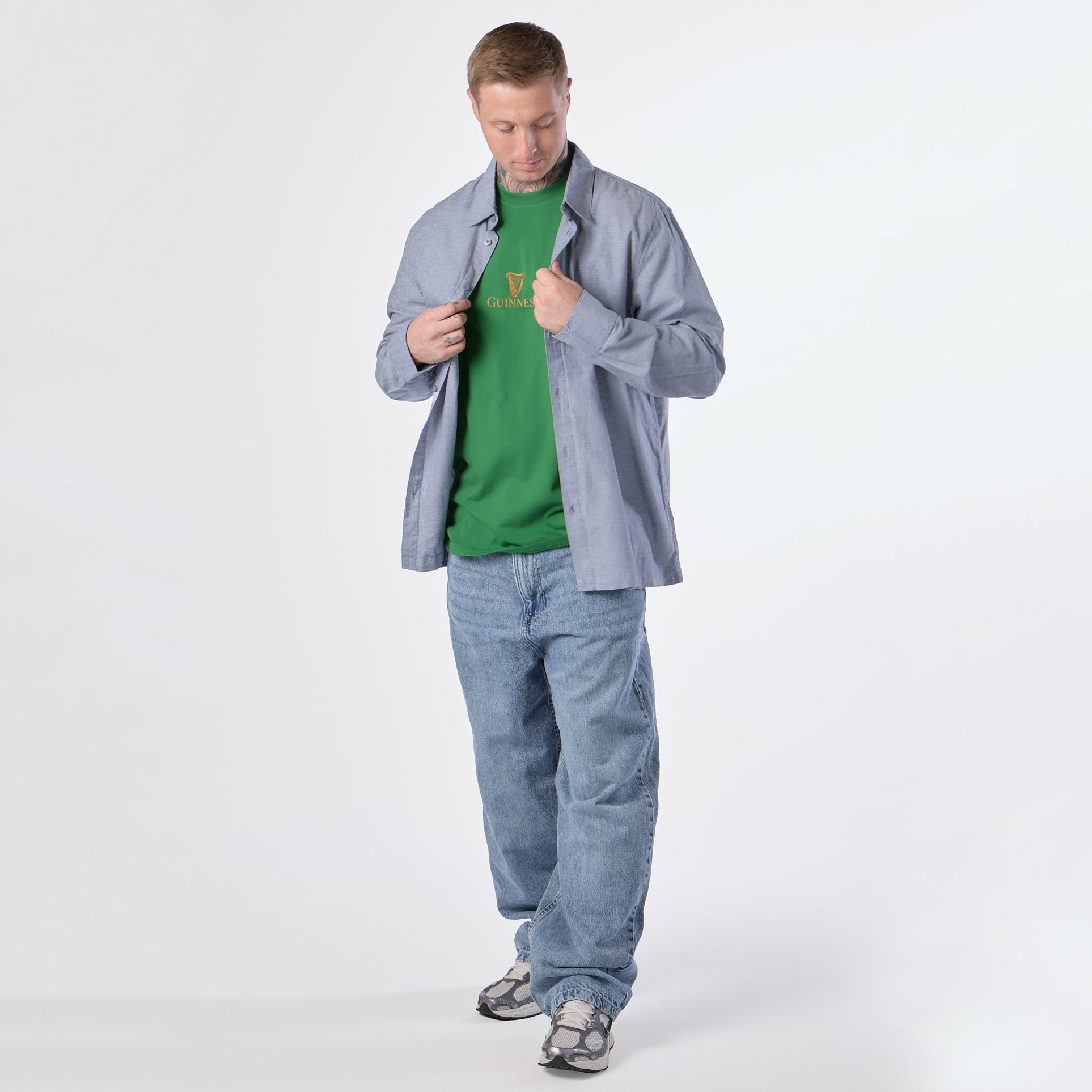 A man stands against a plain white background, wearing the Guinness Gilroy Toucan Tee in green from Guinness Webstore US, an unbuttoned blue shirt, loose blue jeans, and gray sneakers, looking down while adjusting his shirt.